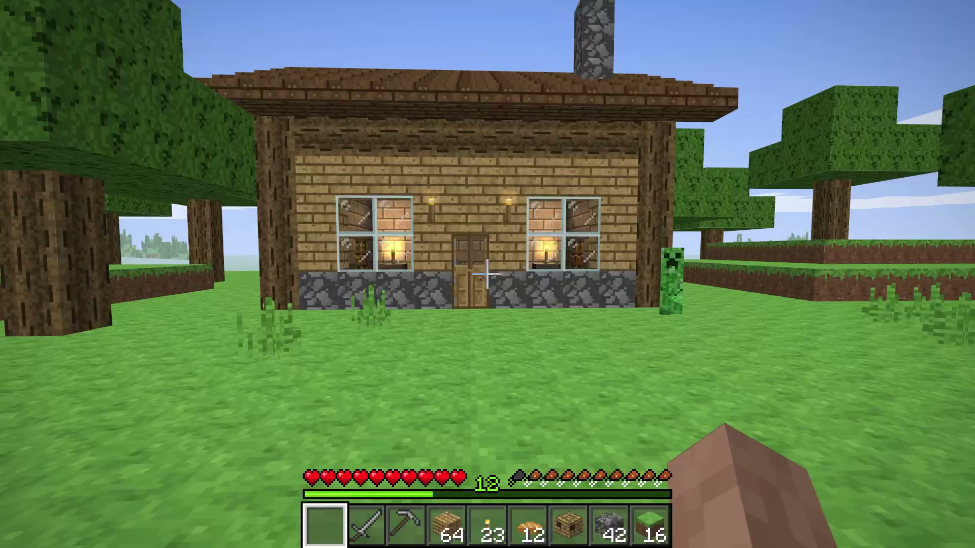 First-person view of a wooden cabin with glowing windows among blocky trees, a creeper on the grass, and a hotbar with hearts along the bottom